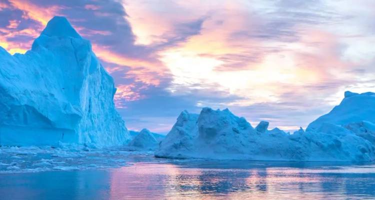 Cielos rosa y naranja brillantes iluminan imponentes témpanos de hielo ártico reflejándose en aguas tranquilas.