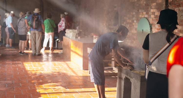 Visitors watch a local craftsman roast grains inside a brick workshop as sunlight streams through smoke.