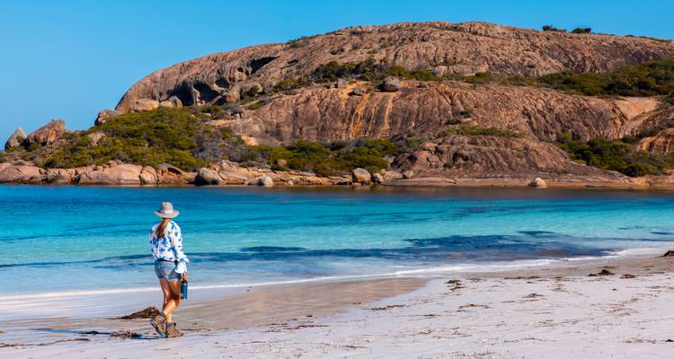 Woman strolls along pristine white-sand beach beside turquoise bay and granite headland.
