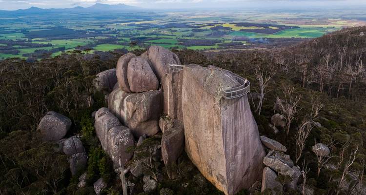 Drone view of massive granite tors with a suspended skywalk platform amid dense forest and patchwork farmland beyond.