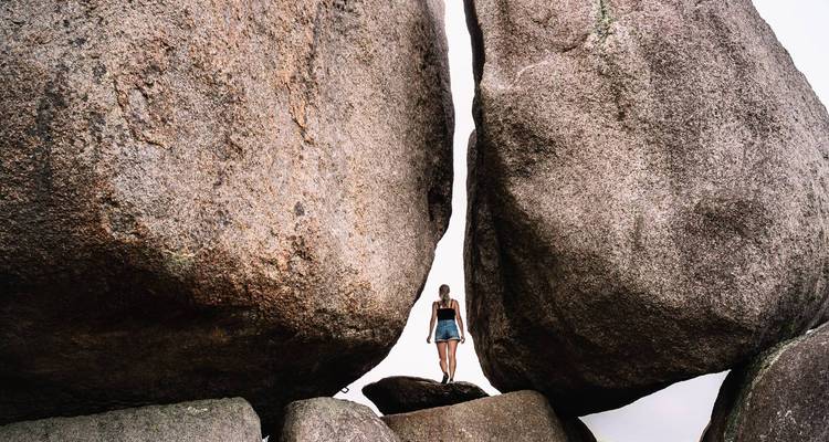 Traveller stands on a ledge between two enormous weathered boulders against a bright sky.