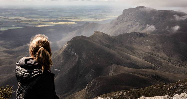 Woman in jacket gazes over misty mountain ridges and a vast valley from a rocky summit.