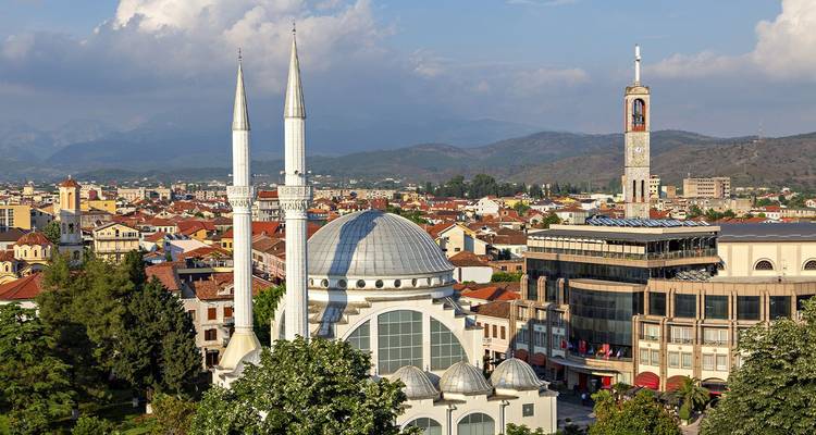 Cityscape featuring a modern white mosque with twin slender minarets, a clock tower and surrounding rooftops set against distant mountains.