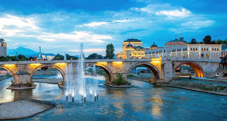 Historic stone bridge with lit arches spanning a river at dusk, fountains rising in front and ornate buildings glowing in the background.