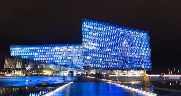 Modern glazen concertgebouw verlicht met blauwe lampen, gereflecteerd op het water 's nachts.