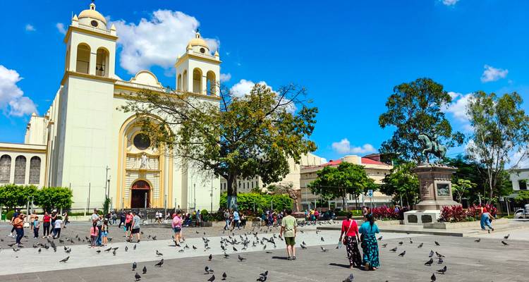Plaza bañada por el sol frente a la Catedral Metropolitana de San Salvador llena de palomas y gente local paseando.