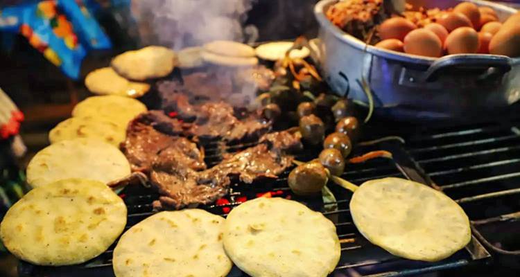 Parrilla de comida callejera cargada de tortillas humeantes, carne chisporroteante, verduras y huevos sobre carbones incandescentes.