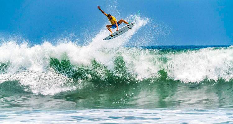 Un surfista lanzándose alto sobre una ola turquesa que se estrella bajo un cielo despejado.