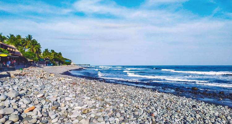 Playa cubierta de guijarros con olas suaves, palmeras junto al mar y cafés en la playa a lo largo de la costa del Pacífico.