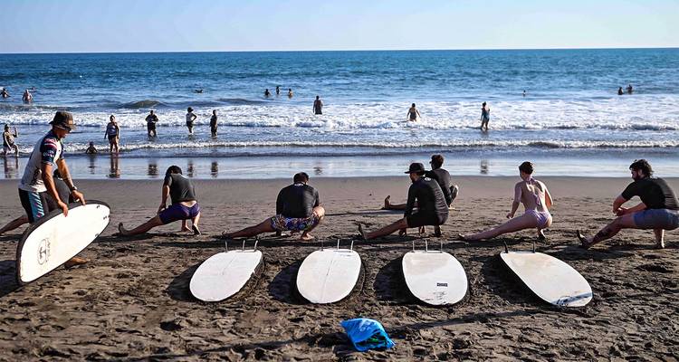 Estudiantes de surf estirándose en la playa arenosa mientras los nadadores disfrutan de las olas que rompen más allá.