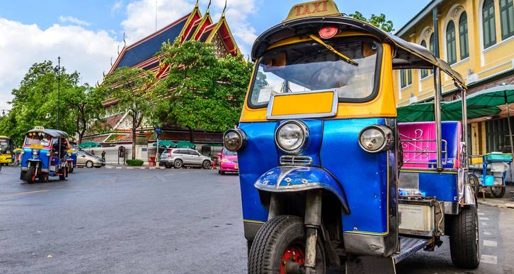 Un tuk-tuk de color azul brillante y amarillo estacionado en una calle de la ciudad con un templo tailandés colorido y un edificio colonial de fondo.