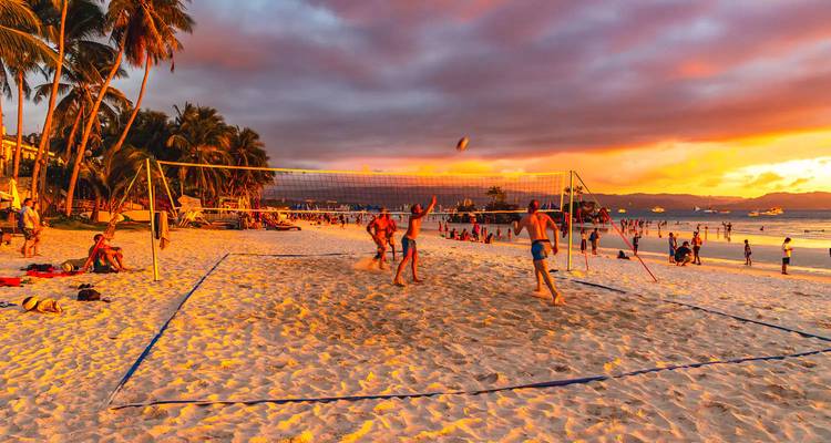 People play beach volleyball on white sand at sunset with orange-pink skies.