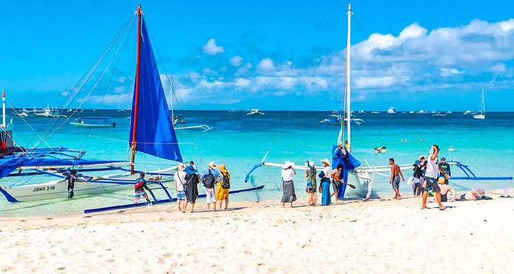 Tourists prepare outrigger sailboats on a bright turquoise beach under clear blue skies.