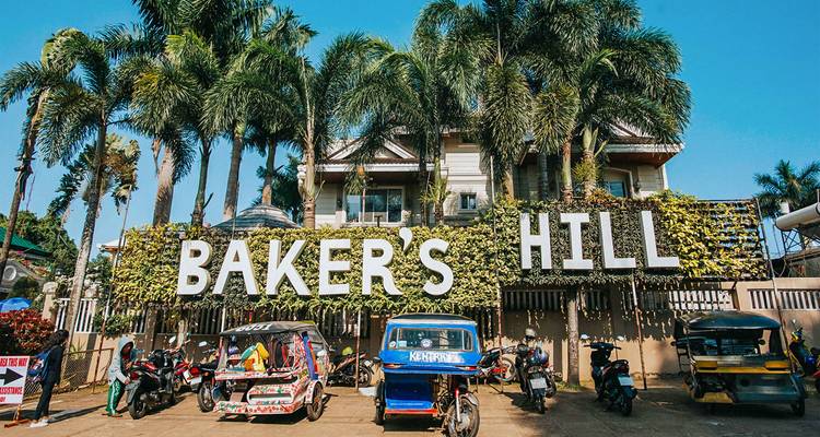 Entrance of Baker's Hill with tall palms and colorful tricycles parked below large sign.