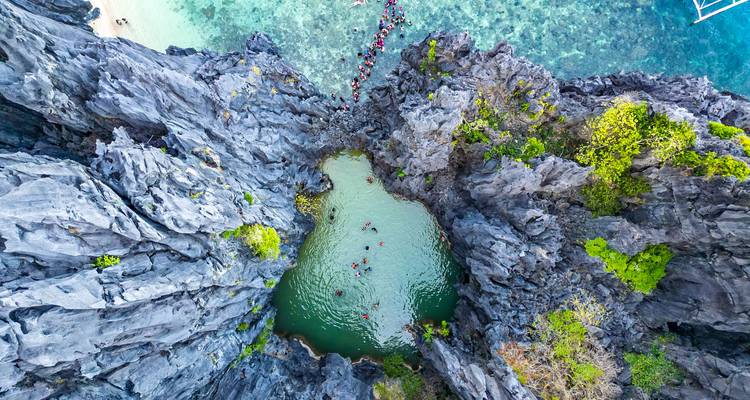 A spectacular aerial view of a small emerald lagoon surrounded by dramatic black limestone cliffs where swimmers form a chain across the water.