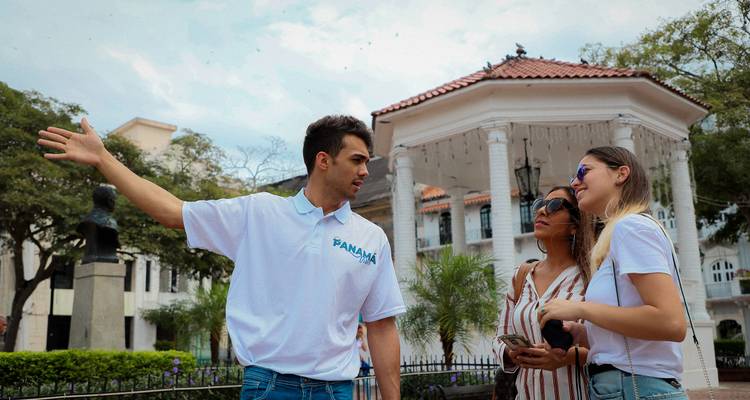 El guía turístico señala hacia la plaza histórica mientras dos mujeres escuchan junto a un quiosco.