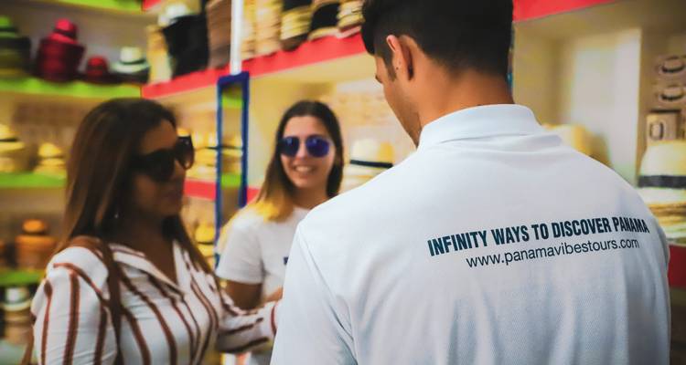Dentro de la tienda de sombreros, un guía con una camiseta con marca habla con dos visitantes sonrientes que usan gafas de sol.