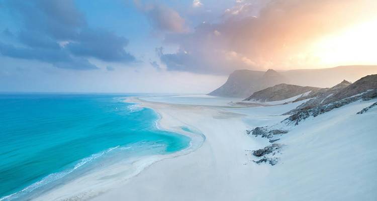 Arena blanca prístina se extiende hacia aguas turquesas junto a un promontorio escarpado bajo un cielo de amanecer resplandeciente.
