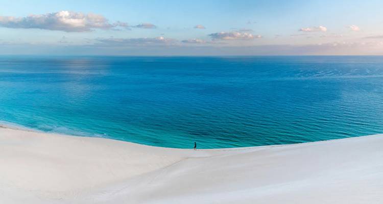 Un viajero solitario se encuentra sobre dunas blancas contemplando el océano azul infinito bajo un suave cielo matutino.