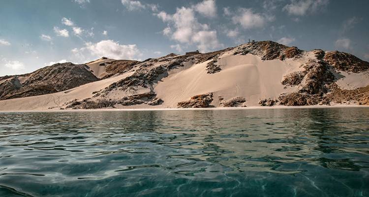 Agua verde azulada clara lame contra una isla rocosa desierta coronada de arena bajo nubes dispersas.