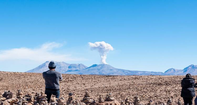 Two travellers watch a distant volcano plume rise above a barren high-altitude plain.