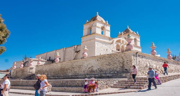 Historic white-stone church with villagers and tourists walking beneath a cloudless sky.