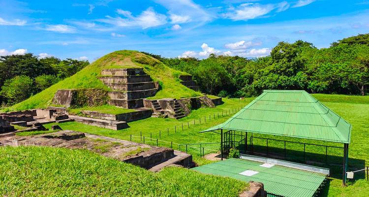 Grassy earthen pyramid and excavated stone foundations at a protected archaeological park