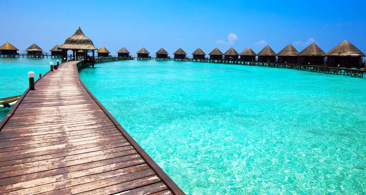 Long wooden boardwalk leads to overwater bungalows above dazzling turquoise lagoon under clear skies.