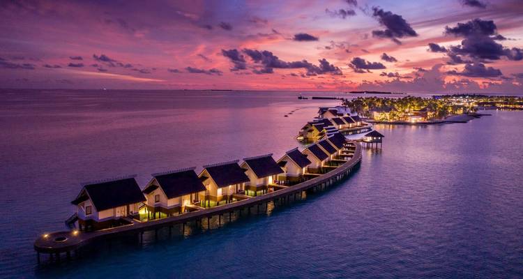 Row of overwater villas curving into calm sea under dramatic pink and purple sunset sky