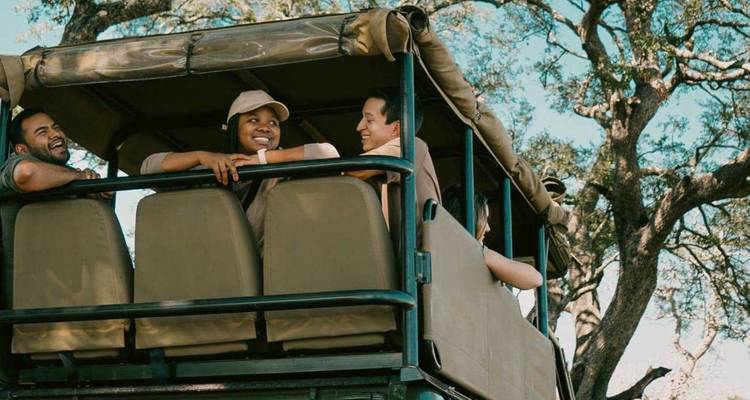 Smiling travellers ride in an open-sided safari vehicle beneath leafy trees on a sunny day.