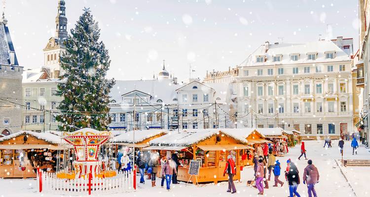 A snowy Christmas market with wooden stalls, a large decorated tree, and historic buildings surrounding Tallinn's town square.