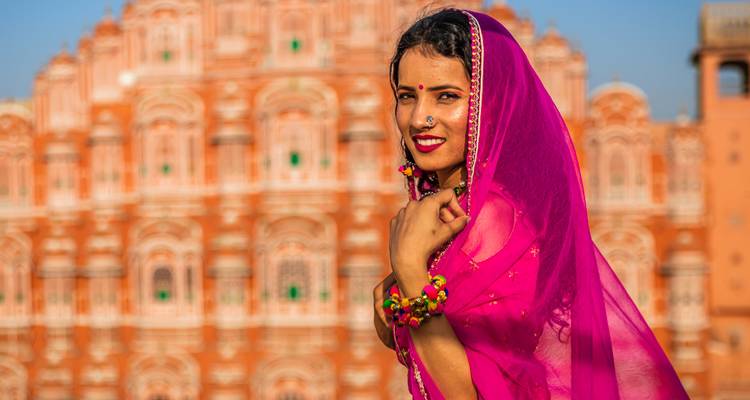 Lächelnde Frau in leuchtend rosafarbenem Sari steht vor der kunstvollen Fassade des Hawa Mahal in Jaipur.