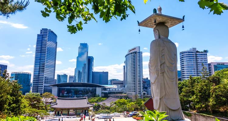 Eine Statue, die eine moderne Stadtsilhouette überblickt, mit traditioneller Architektur im Vordergrund.