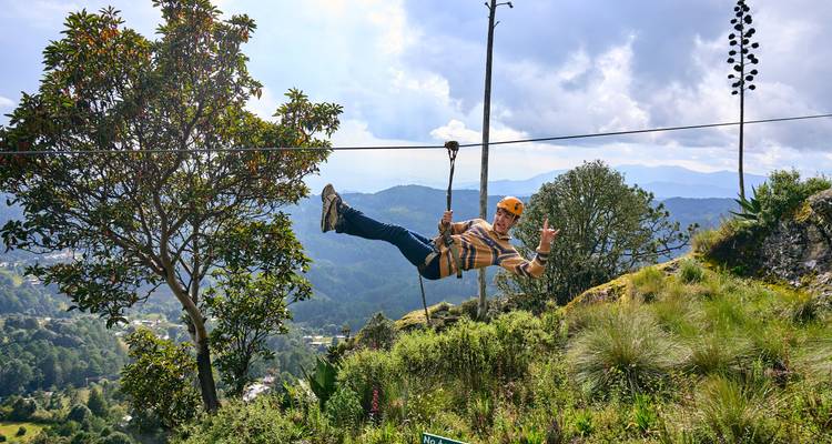 Persona sonriendo y haciendo gestos mientras se desliza rápidamente por un cable sobre un valle pintoresco.