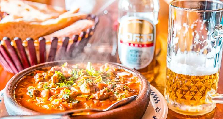 Guiso georgiano abundante servido en un cuenco de barro junto a pan fresco y un vaso helado de cerveza sobre una mesa de madera
