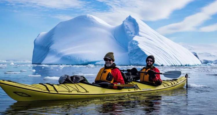 Dos kayakistas en kayaks de mar amarillos remando entre hielo flotante y un iceberg gigante en las tranquilas aguas antárticas.