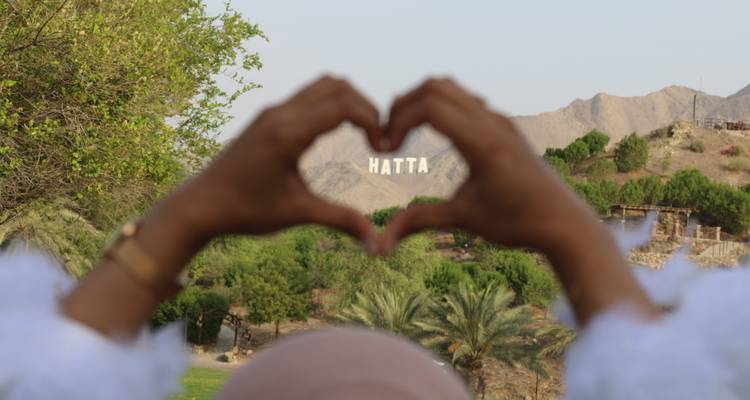 Hände, die eine Herzform bilden und einen Blick auf Berge einrahmen, mit „Hatta" im Hintergrund geschrieben.