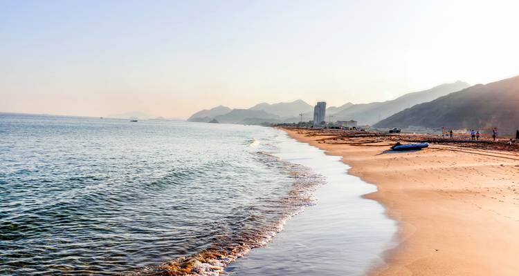 Ein langer Strandküstenabschnitt mit sanften Wellen und fernen Bergen.