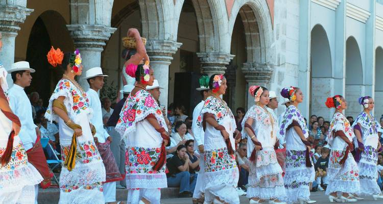 Bailarines yucatecos tradicionales con atuendos bordados de colores vivos actúan frente a arcos coloniales.