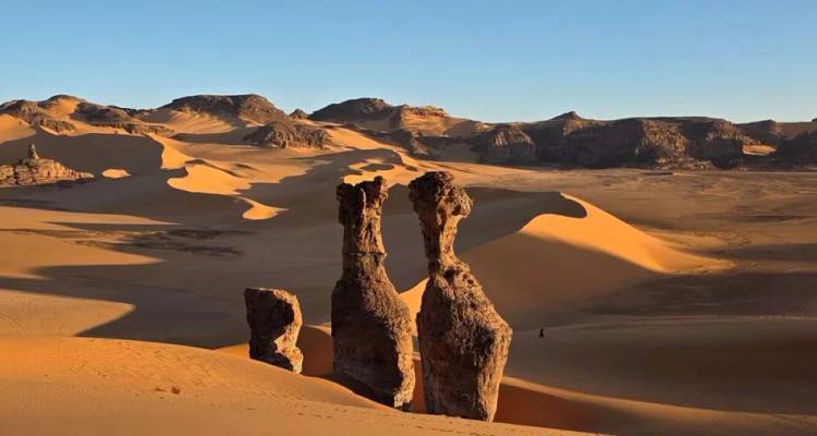 Towering sandstone pillars cast long shadows across the golden dunes of the Sahara Desert.