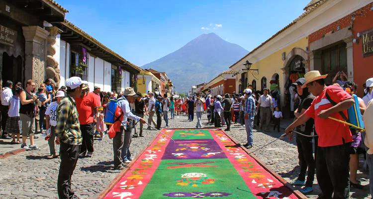 Les foules se pressent dans une rue pavée de galets pour admirer un tapis de sciure coloré lors d'une fête avec un volcan en arrière-plan