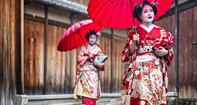 Two elegantly dressed geishas in vibrant kimonos walk with red umbrellas down a wooden lane in light rain.