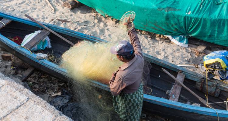 Un pescador repara redes de pesca amarillas junto a una canoa de madera en la arena.