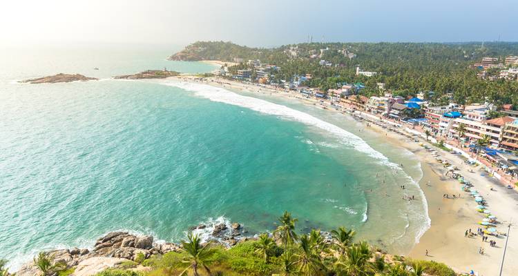Vista aérea amplia de la playa de Kovalam en forma de media luna rodeada de palmeras y la ciudad.