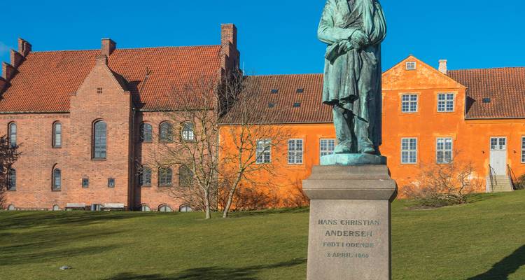 Standbeeld van Hans Christian Andersen staat voor kleurrijke historische gebouwen op een zonnig grasveld.