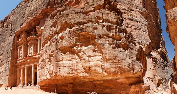 Panoramic rock wall revealing Petra’s iconic Treasury with tiny tourists for scale.