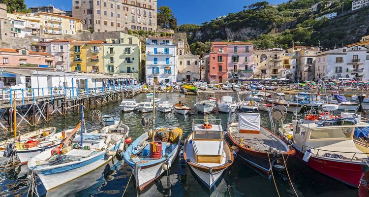 Barcos de pesca coloridos amarrados en un animado puerto italiano rodeado de edificios frente al mar en tonos pastel.