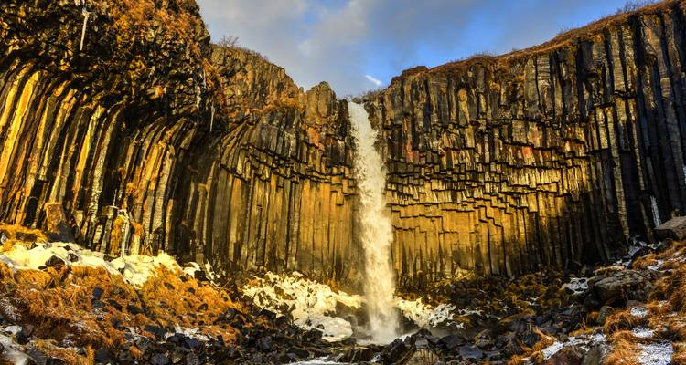 La cascada Svartifoss se precipita sobre columnas de basalto oscuro cubiertas de nieve bajo una luz dorada.