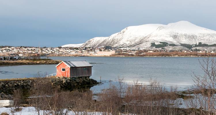 Rotes Schuppen am felsigen Fjordufer mit schneebedeckten Bergen und Stadt in der Ferne.