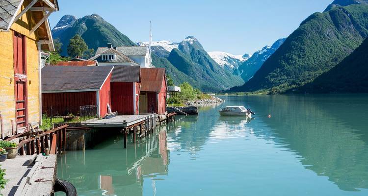 Kleines Dorf an der Fjordküste mit roten Bootshäusern, die sich im türkisfarbenen Wasser unter schneebedeckten Gipfeln spiegeln.